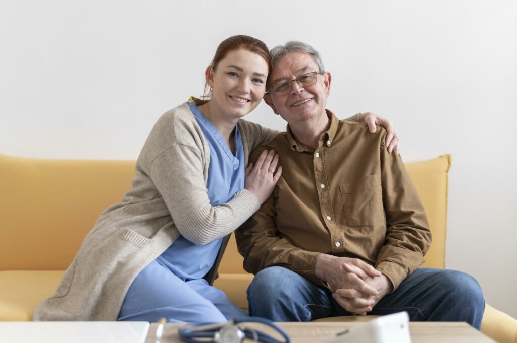 A female care nurse and elderly man smiling towards the camera. The nurse has her arm around the man's back and hand on his shoulder.