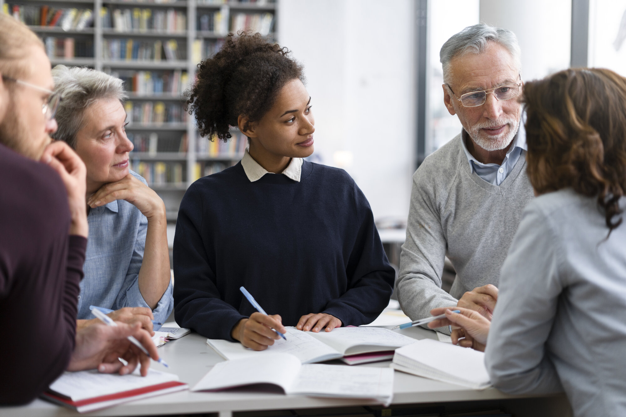 A group of mixed young and senior people are sitting together taking notes. Three of them are looking over at a man to the right side of the photo talking to a brunette woman sitting across from him.