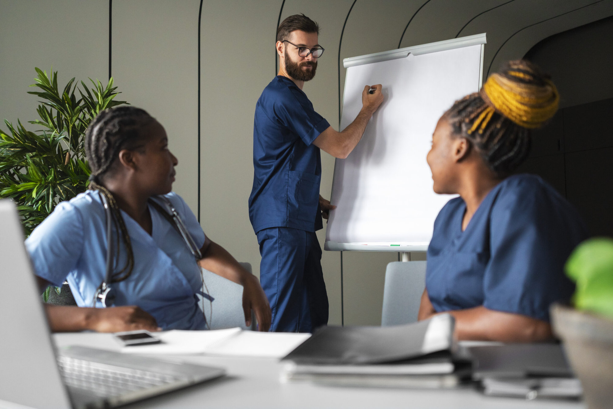 Two female nurses are sitting and looking at their male colleague who is standing between them from a distance, he is preparing to write on a whiteboard.