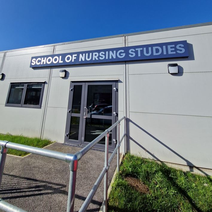 A silver rectangular building with a navy blue sign that reads 'School of Nursing Studies' across the top. There is a metal railing that makes a path for people to follow to reach the door.