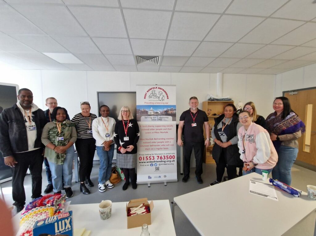 A group of adult students standing and smiling towards the camera. In the centre of the picture is a pull-up banner which the two teachers are standing and smiling next to.