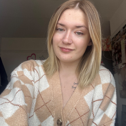 A blonde female teenager smiling at a camera. She is wearing a white cardigan with peach coloured diamond patterns